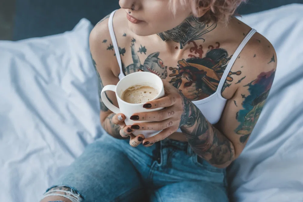 Chica con tatuajes en el cuerpo tomando un café sentada en la cama. La personalidad de los tatuajes
